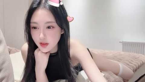 Media: Video of an East Asian woman with long black hair, wearing a black dress, lying on a beige bed in a minimalist room.