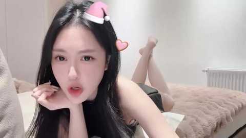 Media: Video of an East Asian woman with long black hair, wearing a pink Santa hat and black lingerie, lying on her stomach on a bed, legs crossed, in a minimalist bedroom.