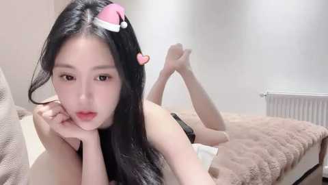 Media: Video of an East Asian woman with long black hair and fair skin, wearing a Santa hat, lying on her stomach on a bed in a minimalist, softly lit room.