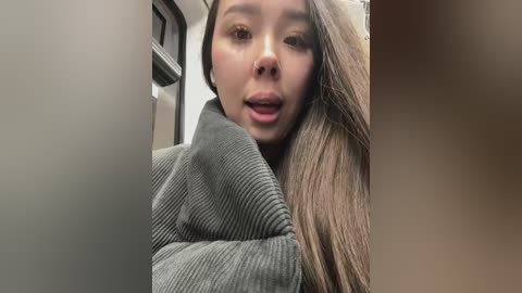 Media: Video of a young Asian woman with straight, light brown hair, wearing a dark green knitted sweater, inside a dimly lit bus. She has a neutral expression and is slightly out of focus.