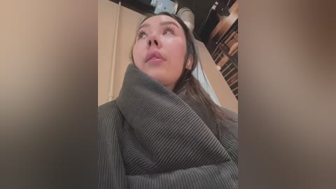 Media: Video of a young woman with straight, dark hair, wearing a gray, ribbed sweater, looking upward, against a blurred background of wooden shelves.