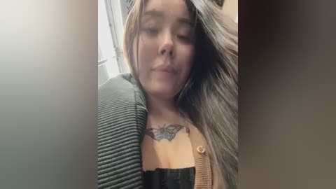 Media: Video of a light-skinned woman with long, straight, light brown hair, wearing a gray cardigan over a black bra. She has a butterfly tattoo on her chest. The background is blurry.