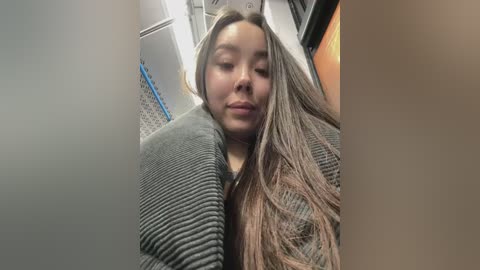 Media: A video of a young Asian woman with long, straight brown hair and fair skin, wearing a gray knitted sweater, sitting in a train with metal railings and gray walls in the background.