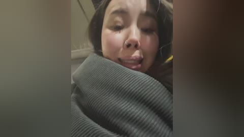 Media: Video of a young Asian woman with dark hair, closed eyes, and slightly open mouth, wrapped in a gray blanket, indoors.