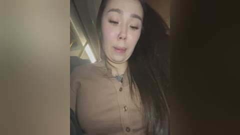 Media: Video of an Asian woman with long black hair, wearing a beige cardigan and a turquoise necklace, eyes closed, seated in a dimly lit car interior.