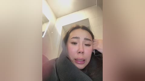 Media: A blurry, low-resolution video of a young Asian woman with long hair, wearing a grey sweater, looking upset and holding her face in a bathroom with beige tiles and fluorescent lighting.