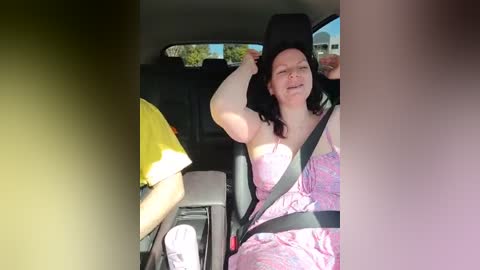 Media: Video of a woman in a car, wearing a pink dress, black seatbelt, yellow towel, black headrest, smiling, sunlit interior, relaxed pose.