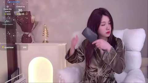 Media: Video of an Asian woman with long black hair, wearing a green satin robe, using a hair dryer in a modern, minimalist bedroom with white furniture and soft lighting.