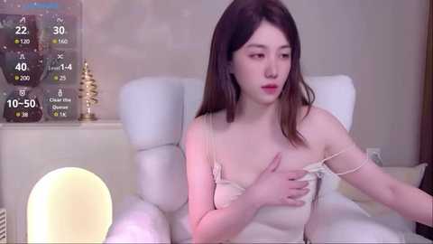 Media: Video of an Asian woman with light skin, long dark hair, wearing a white strapless dress, covering her chest with her hands, sitting on a white chair in a cozy room with a glowing orb lamp, thermometer, and Christmas decorations.