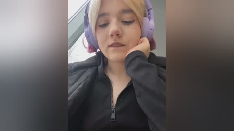 Media: A video of a young woman with light skin and short blonde hair, wearing a black hoodie, resting her head on her hand, listening to music with large purple headphones.