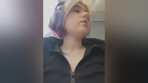 Media: A video of a young, fair-skinned person with shoulder-length, pastel-colored hair, wearing a black hoodie and purple headphones. The background is blurry, possibly indoors.