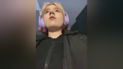 Media: Video of a young woman with light skin, blonde hair, and purple headphones, wearing a black hoodie, viewed from below in a dimly lit room.