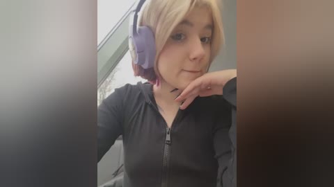 Media: Video of a young woman with fair skin and short blonde hair, wearing purple headphones and a black hoodie, resting her chin on her hand, indoors with blurred background.