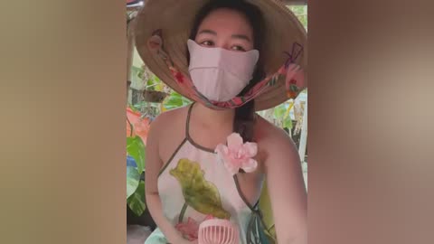 Media: Video of an Asian woman with a wide-brimmed straw hat, face mask, and floral-patterned top, holding a pink hairbrush, standing outdoors in a garden.