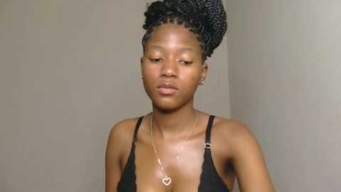 Media: Video of a young, dark-skinned woman with braided hair, wearing a black spaghetti strap top, and a gold necklace with a heart pendant. She has a neutral expression against a plain beige wall.