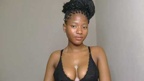 Video of a young Black woman with medium brown skin, braided hair in a high bun, wearing a black lace bra and a heart pendant necklace, standing against a plain beige wall.