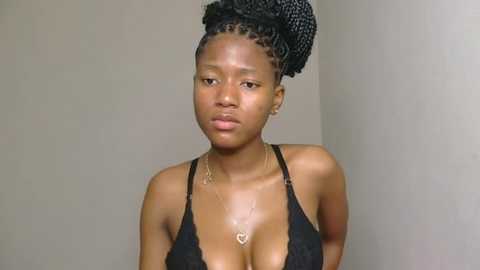 Media: Video of a young Black woman with a slender physique, wearing a black lace bra, her hair styled in intricate braids, set against a plain beige wall background.