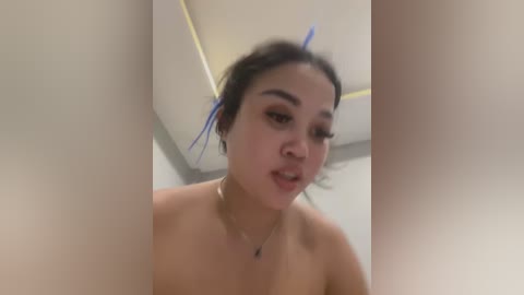 Media: Video of a young Asian woman with medium skin tone, black hair styled with blue ribbons, wearing a nose ring, and a small silver necklace. She is topless, indoors with a white ceiling.