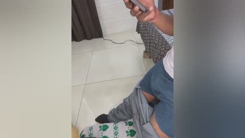 Media: Video of a person in a living room, wearing a gray t-shirt, blue shorts, and black socks, with a gray sweater partially pulled down, holding a smartphone, sitting on a white tiled floor.