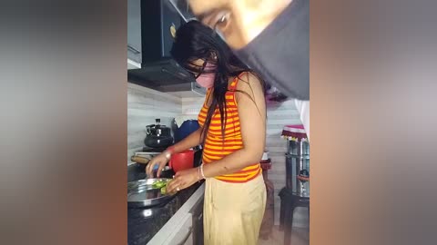 Media: Video of a young woman with long black hair, wearing a striped orange and yellow shirt and beige pants, cooking in a modern kitchen with white tiles, black countertops, and a black kettle.