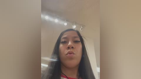 Media: Video of a young Asian woman with long black hair, light brown skin, and a red shirt, making a pouty face in a well-lit room with beige walls and overhead lights.