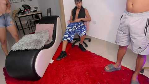 Media: Video of a muscular, shirtless man with blue Adidas shorts sitting on a black and white chaise lounge, surrounded by three shirtless men in casual shorts on a red shag rug in a minimalistic, modern room.