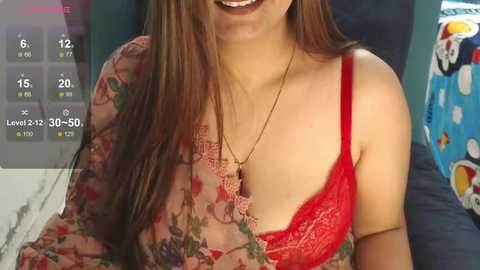 Media: Video of a smiling woman with long brown hair, wearing a floral dress with a red lace bralette, seated against a blue cushion.