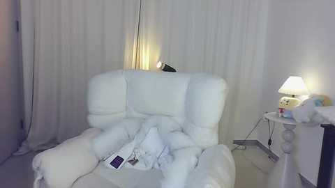 Media: A video of a cozy, dimly lit room featuring a white armchair, covered with white blankets, a nightstand with a lamp, and sheer white curtains in the background.