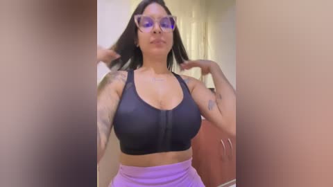 Video of a light-skinned woman with straight black hair, wearing a black sports bra and purple high-waisted leggings, adjusting her hair in front of a beige wall and brown cabinet.