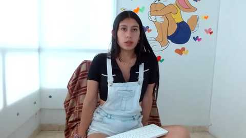 Media: Video of a young Latina woman with long black hair, wearing overalls and a black shirt, sitting on a plaid couch, holding a keyboard. Background features a colorful Looney Tunes mural.