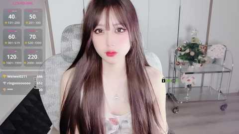 Media: A video of a young Asian woman with long, straight brown hair, wearing a white floral top and red lipstick, sitting on a gray chair in a minimalistic room.