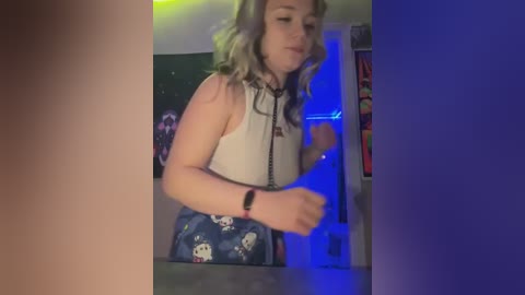 Media: Video of a young woman with wavy, light brown hair, wearing a sleeveless white top and blue skirt with Hello Kitty pattern. She stands in a dimly lit room with green and blue lighting, holding a phone.