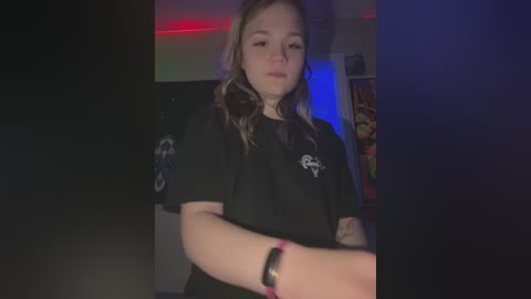 Media: Video of a young, fair-skinned woman with wavy blonde hair, wearing a black t-shirt with white logo, standing indoors with dim lighting, colorful wall art, and red LED strip lights.