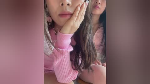 Media: Video of two young women with long hair, one with a pink sweater, the other with a light pink top. They're in a dimly lit room, looking at the camera.