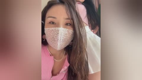 Media: Video of an Asian woman with long brown hair wearing a pink ribbed sweater, a face mask, and pearl earrings, seated with a child partially visible behind her.