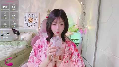 Media: Video of an Asian woman with fair skin, long black hair, wearing a pink floral robe, drinking tea, in a cozy bedroom with floral wallpaper, a bed, and fairy lights.