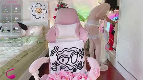 Media: A video shows a young woman in white lingerie adjusting her bra, standing beside a pink gaming chair with a cartoon face. The room is brightly lit, with a bed and wall decor in the background.