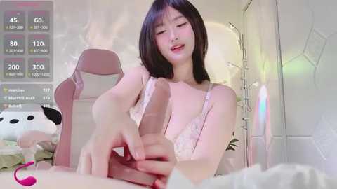 Media: Video of an Asian woman with straight black hair, wearing a white lace bra, sitting on a pink gaming chair, stroking a large, erect penis. Background includes a wall, fairy lights, and plush toys.