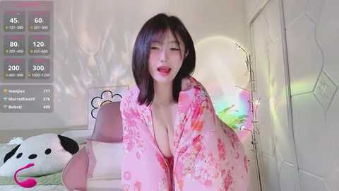 Media: Video of an Asian woman with straight black hair, wearing a pink floral kimono, sitting on a bed in a bright, minimalistic room with white walls and a panda plush toy.