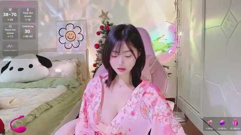 Media: A young East Asian woman with dark hair and fair skin sits on a bed in a cozy, festive bedroom, wearing a pink floral kimono, with a Christmas tree and presents in the background.