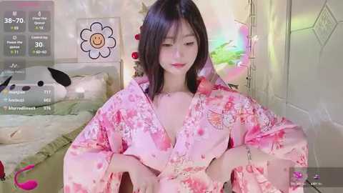 Media: Video of a young Asian woman with straight black hair, wearing a pink floral kimono, sitting in a cozy bedroom with a plush toy and a colorful, star-shaped light in the background.