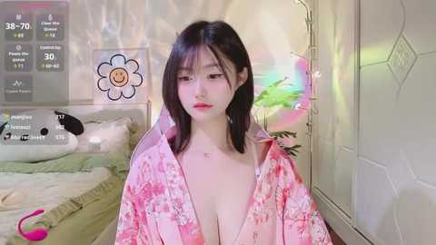 Media: A video of an East Asian woman with fair skin, straight black hair, and a pink floral kimono, sitting in a cozy bedroom with a green blanket, white walls, and a glowing pink light.