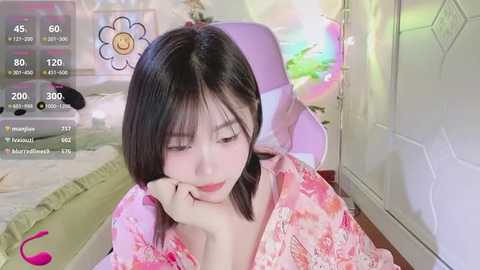 Media: Video of a young Asian woman with straight black hair, wearing a pink floral kimono, resting her chin on her hand, in a cozy, softly lit room with a pink chair and green blanket in the background.