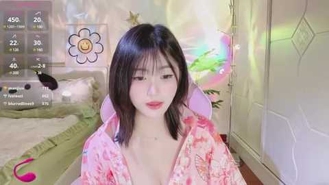 Media: A video of an East Asian woman with shoulder-length black hair, fair skin, and red lipstick, wearing a pink floral robe, sitting in a pastel bedroom with a bed, fairy lights, and a large flower wall art.