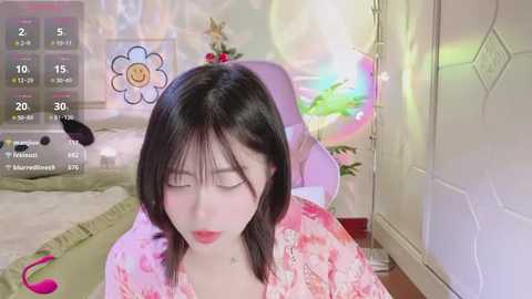 Media: A video of a young Asian woman with straight black hair, wearing a pink floral dress, sitting in a pastel-colored room with a large, colorful flower wall decoration and a green blanket.