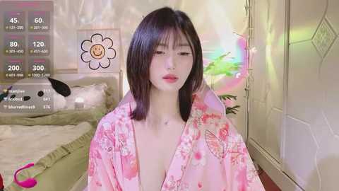 Media: A video of an Asian woman with shoulder-length black hair, wearing a pink floral robe, sitting in a dimly lit bedroom with a bed, flower wall art, and a pink toy on the floor.