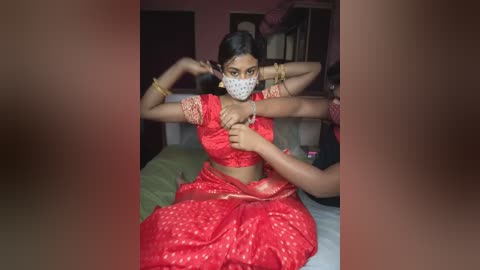 Media: Video of a young woman with dark skin and medium build, wearing a red sari with gold embroidery, and a white polka-dot mask, being restrained by two men in a dimly lit bedroom.