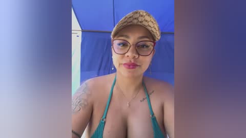 Media: Video of a plus-sized, light-skinned Latina woman with a large chest, wearing a teal bikini top, a beanie, and glasses, inside a blue-tinted, curtained tent.