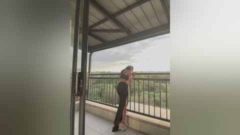 Video of a woman with long hair, wearing a black top and pants, standing on a balcony with a green landscape view, holding her phone, under a cloudy sky.