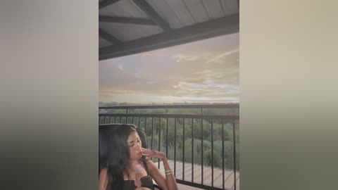 Media: A video of a young woman with long black hair, wearing a black top, sitting pensively on a balcony with a serene sunset view over a vast green landscape.